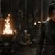 The Devil's Secretary 5 A stern woman in a dark, textured suit staring intensely, standing in a dim, gothic stone chamber. To her left burns a bright fire in an ornate, horned skull brazier, with skeletons and candles visible on the stone steps in the shadowy background from The Devil's Secretary film